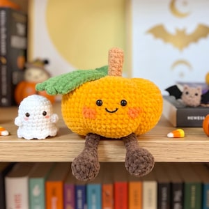 May include: A crocheted orange pumpkin plush with a green leaf, a tan stem, and dangling brown legs. The pumpkin has a smiling face with black eyes and pink cheeks. Beside it sits a small white crocheted ghost plush with black eyes and pink cheeks.