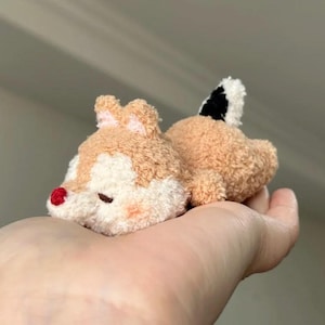 Op de afbeelding: Een kleine, pluche eekhoornknuffel in beige en crèmekleur met een pluizige textuur. Het speelgoed heeft een piepkleine rode neus, kleine oortjes en een korte staart met een opvallende zwart-witte vlek. Het heeft een ronde, compacte vorm.
