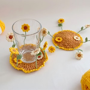 May include: Two round, crocheted coasters in mustard yellow with scalloped edges. Each coaster features attached, three-dimensional crocheted flowers in white and yellow with green stems. A clear glass sits on one coaster, surrounded by the flowers.