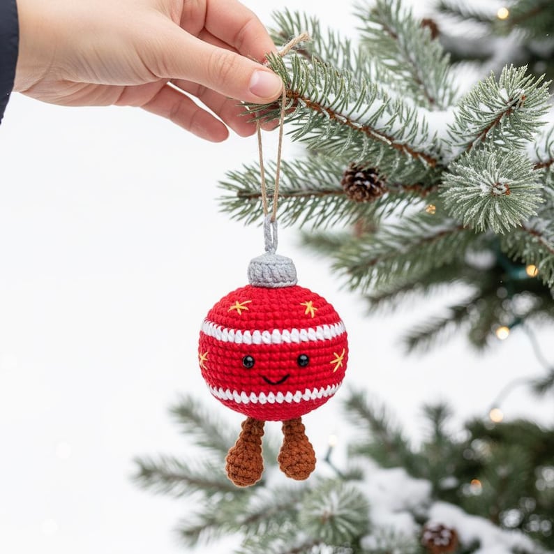 Peut inclure: Une boule de No&euml;l rouge et blanche crochet&eacute;e, avec un visage souriant et des jambes marron. L'ornement est d&eacute;cor&eacute; de rayures blanches et d'&eacute;toiles jaunes. Il est suspendu &agrave; une ficelle en ficelle naturelle.