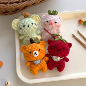 Op de afbeelding: Vier kleine, handgemaakte gehaakte knuffels in de vorm van dieren met fruit-thema accessoires. De set bevat een lichtgroene muis, een lichtroze varken, een oranje beer en een donkerrode kat, elk met geborduurde details op het gezicht.