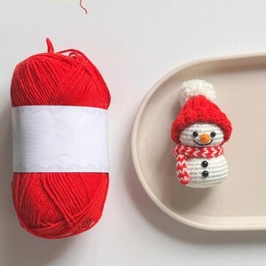 May include: A red yarn ball with a blank white label sits next to a crocheted snowman figurine. The snowman wears a red hat and scarf, with black button eyes and a carrot nose. The snowman is on a beige tray.