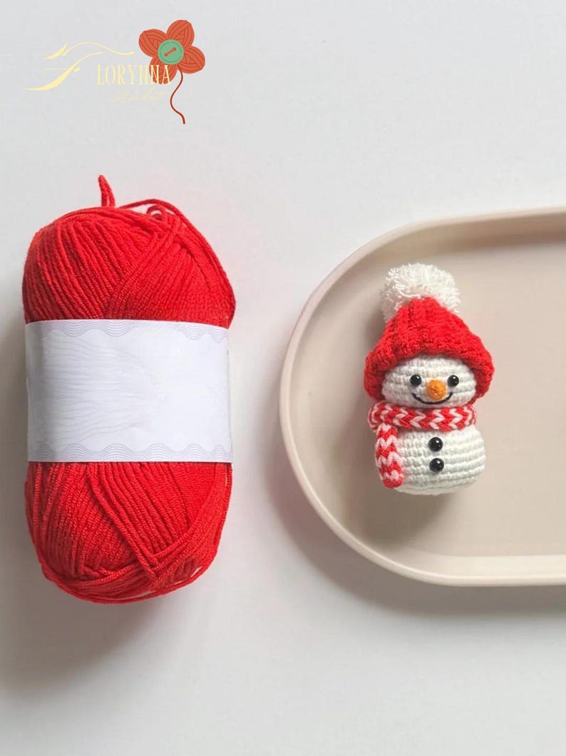 May include: A red yarn ball with a blank white label sits next to a crocheted snowman figurine. The snowman wears a red hat and scarf, with black button eyes and a carrot nose. The snowman is on a beige tray.