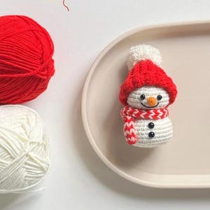 May include: A handmade crochet snowman with a red hat, scarf, and black button eyes. A ball of red yarn and a ball of white yarn are in the background. The snowman is on a light pink oval plate.