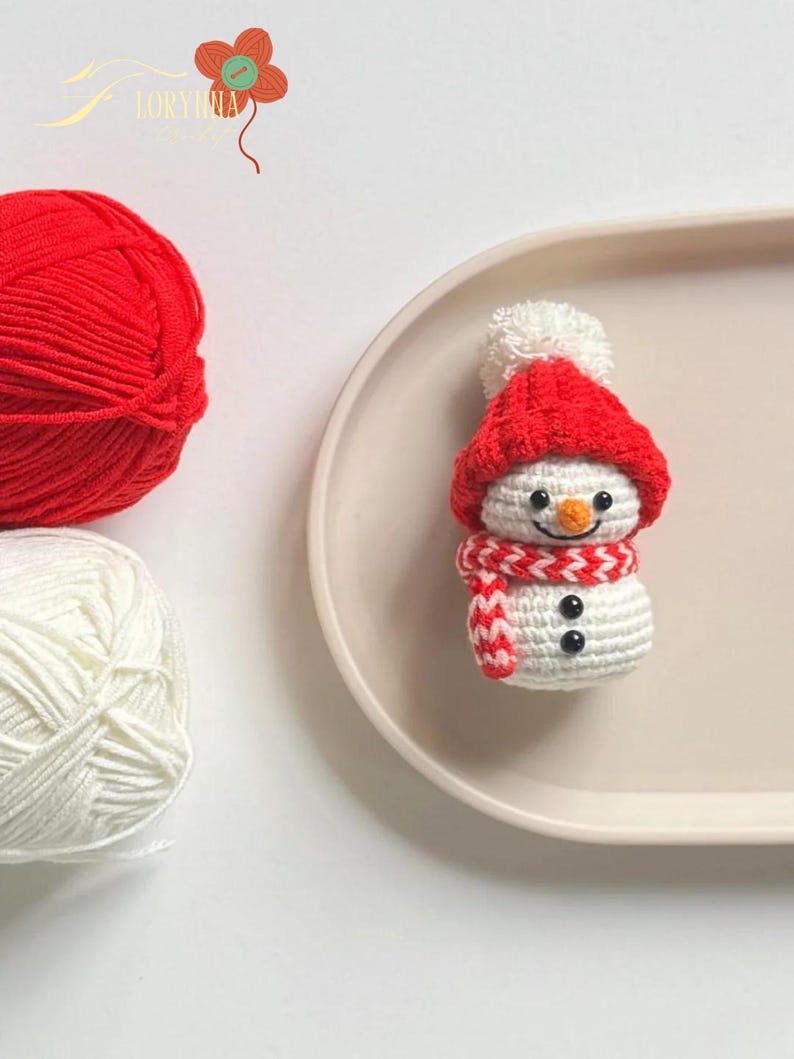 May include: A handmade crochet snowman with a red hat, scarf, and black button eyes. A ball of red yarn and a ball of white yarn are in the background. The snowman is on a light pink oval plate.