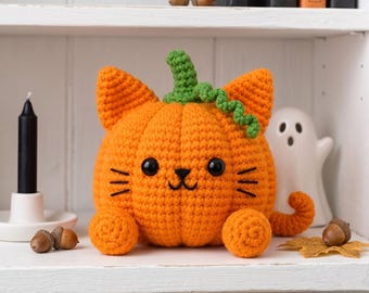 May include: A crocheted orange pumpkin shaped like a cat with a green stem, black plastic eyes, and embroidered whiskers. The plush figure features two pointed ears, two round front paws, and a curled tail, sitting on a white shelf surface.