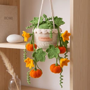 May include: A hanging crochet planter featuring a cream-colored pot with a scalloped edge and a leather tag labeled HANDMADE. The pot is adorned with crocheted green vines, curly tendrils, large green leaves, bright yellow flowers, and orange pumpkins.
