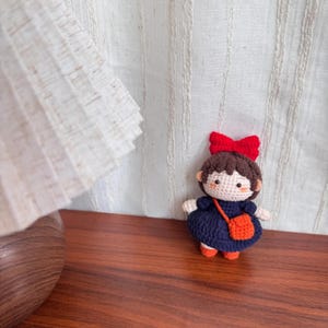 May include: A handmade crochet doll with brown hair and a red bow. The doll wears a navy blue dress and an orange purse. The doll has orange shoes and is standing on a wooden surface.