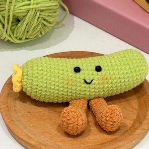 Puede incluir: Un juguete de ganchillo hecho a mano con forma de calabacín verde con una cara sonriente y piernas naranjas. El juguete está colocado sobre un plato de madera, con una madeja de hilo verde en el fondo.