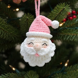 May include: A crocheted Santa Claus Christmas decoration. The ornament has a pink hat with a white bobble, a white beard, and a smiling face with pink cheeks. It is suspended from a Christmas tree.