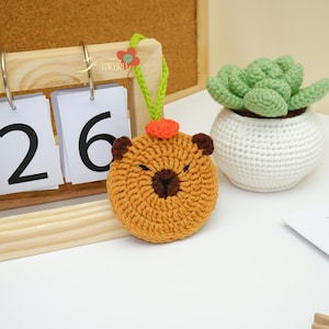Könnte beinhalten: Gehäkeltes Capybara-Ornament in Brauntönen, mit dunkelbraunen Akzenten für Augen und Nase. Es hat kleine, abgerundete Ohren und eine kleine orangefarbene Blume oben. Eine grüne Schlaufe ist zum Aufhängen befestigt. Eine gehäkelte Sukkulente in einem weißen Topf ist ebenfalls sichtbar.