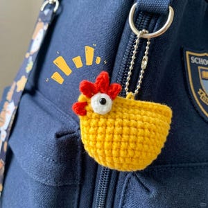 May include: A yellow crocheted chicken keychain with a red comb and a white eye patch featuring a black pupil. The chicken is attached to a silver-toned metal ball chain. Above the chicken, there are yellow graphic lines indicating movement.