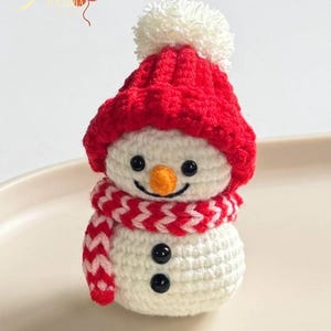 May include: A handmade crochet snowman with a red hat and scarf. The snowman has a white body, black button eyes, and a carrot nose. The red hat has a white pom-pom, and the scarf is red and white. A festive winter decoration.