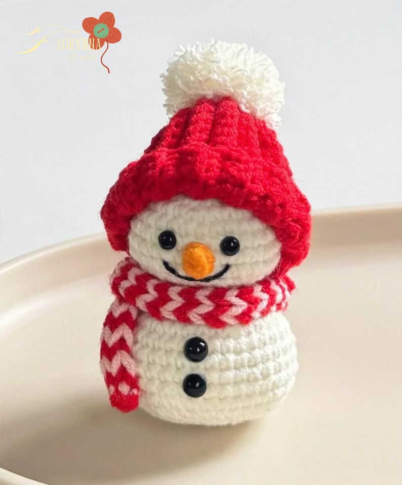 May include: A handmade crochet snowman with a red hat and scarf. The snowman has a white body, black button eyes, and a carrot nose. The red hat has a white pom-pom, and the scarf is red and white. A festive winter decoration.