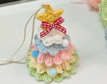May include: A handmade, crocheted Christmas tree ornament. The tree is multi-colored, with layers of pastel colors, a yellow star on top, and a red and white checkered bow. Small pom-poms and a white string are attached for hanging.