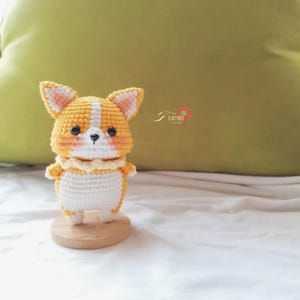 May include: A crocheted corgi dog figurine in shades of orange, white, and peach, standing on a small wooden base. The handmade amigurumi features a white chest, a frilly collar, and black bead eyes. The ears and face have peach accents.