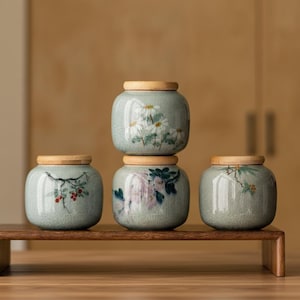 Op de afbeelding: Vier lichtgroene keramische voorraadpotten met een craquelé glazuurafwerking en houten deksels. Elke pot is voorzien van handgeschilderde botanische illustraties, waaronder witte madeliefjes, roze bloemen en rode bessen aan takken, gestapeld.