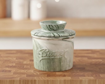 May include: A ceramic butter crock featuring a white and sage green marbled pattern. The cylindrical container has a matching lid with a knob handle and the word Butter embossed in cursive script on the front side of the main body.