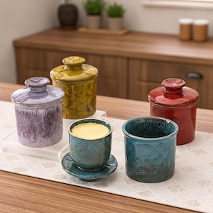 May include: A collection of ceramic butter crocks in various colors including lavender, mustard yellow, red, and blue. Each crock features a cylindrical base with a matching lid, while one blue crock is shown open with a block of yellow butter inside.