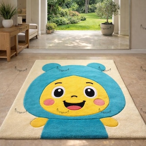 Könnte beinhalten: Rechteckiger Teppich mit einem Cartoon-Charakter-Design. Der Teppich hat einen cremefarbenen Hintergrund mit einem großen, lächelnden gelben Gesicht, das eine blaue Kapuze trägt. Die Figur hat große schwarze Augen und rosa Wangen. Der Teppich liegt auf einem gefliesten Boden.