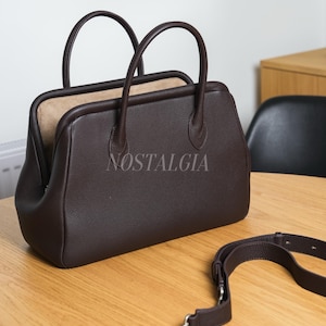 May include: A brown leather handbag with two top handles and a detachable shoulder strap. The bag has a rectangular shape with rounded edges and a textured surface. The word "NOSTALGIA" is printed on the front of the bag.