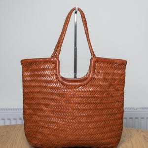 May include: A medium brown woven leather tote bag featuring a herringbone pattern throughout the body. The bag has two braided leather handles and a U-shaped opening at the top centre. The text SchatzSteinbach is visible in white on the bag.