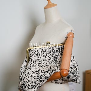 Peut inclure: Une pochette avec un motif tacheté noir et blanc, tenue par un mannequin en bois. La pochette a un cadre et un fermoir de couleur dorée. Le mot "NOSTALGIA" est visible sur la pochette.