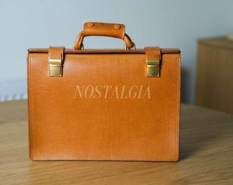 May include: A tan leather briefcase with gold-colored clasps and handle. The word "NOSTALGIA" is printed on the front. The briefcase is sitting on a wooden table.