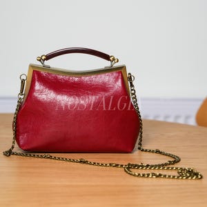 May include: A red handbag with a gold-colored frame and chain strap. The bag has a dark brown handle and the word "NOSTALGIA" is printed on the front. The bag is sitting on a wooden table.