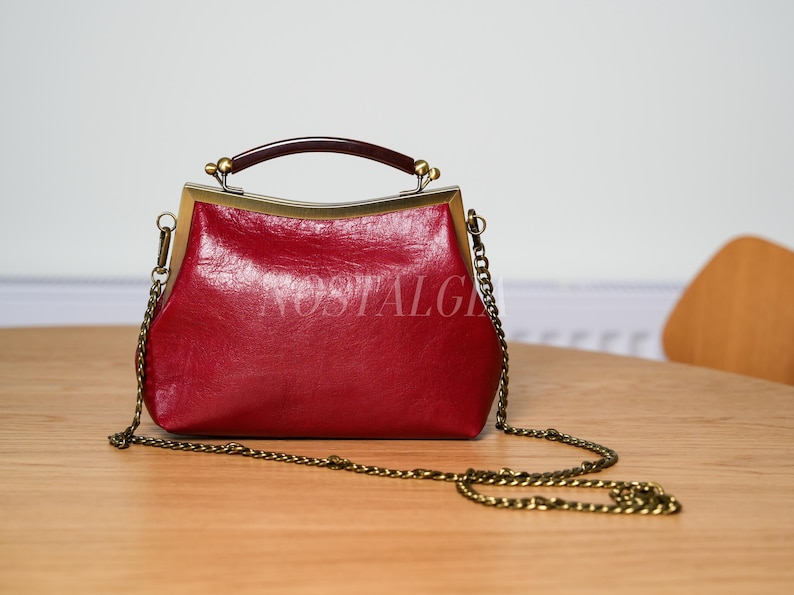 May include: A red handbag with a gold-colored frame and chain strap. The bag has a dark brown handle and the word "NOSTALGIA" is printed on the front. The bag is sitting on a wooden table.