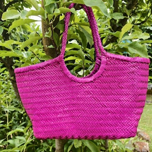 May include: A vibrant magenta tote bag featuring a woven herringbone pattern throughout the body. The bag has two integrated braided handles that extend from the top opening, creating a structured silhouette with a wide base and tapered sides.