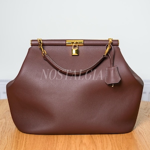 May include: A brown leather handbag with a gold-colored clasp and chain straps. The bag has a structured shape with a top handle and a small tag. The word "NOSTALGIA" is printed on the bag.