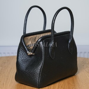 May include: Black leather handbag with two curved handles. The bag has a textured surface and a silver-colored clasp. The interior of the bag is a light tan color. The bag is sitting on a wooden surface.
