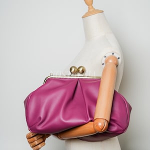 Può includere: Una pochette color magenta con una consistenza morbida, arricciata e plissettata. La borsa presenta una chiusura a telaio in metallo dorato sormontata da due grandi pomelli sferici dorati. Il materiale ha una finitura liscia, opaca e forma rotonda.