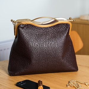 Könnte beinhalten: Eine dunkelbraune Lederhandtasche mit strukturierter Oberfläche und goldfarbenem Verschluss. Die Tasche hat einen geschwungenen Griff und eine rechteckige Form. Ein schwarzes Smartphone und ein Lippenstift liegen auf der Holzoberfläche.