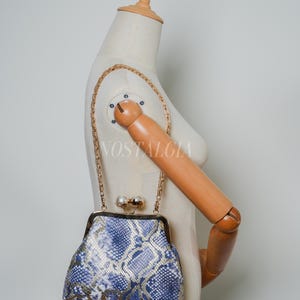 Op de afbeelding: Een handtas in vintage-stijl met een blauw en goud slangenpatroon. De tas heeft een gouden kettingriem en een sluiting met een parelaccent. Het woord "NOSTALGIA" is zichtbaar op de etalagepop.