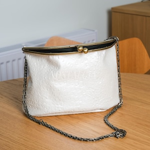 May include: A white, textured clutch bag with a metal chain strap. The bag has a clasp closure with gold-coloured accents. The bag is on a wooden surface.