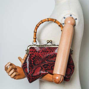 Peut inclure: Un sac à main rouge et noir à motif peau de serpent avec une poignée en bambou. Le sac a un fermoir en métal et une forme structurée. La poignée est incurvée et en bambou brun clair.
