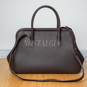 May include: A brown leather handbag with a top handle and a detachable shoulder strap. The bag has a rectangular shape with rounded corners and the word "NOSTALGIA" is printed on the front. The bag is sitting on a wooden table.