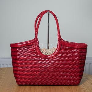 May include: A deep red woven tote bag featuring a herringbone pattern, two braided handles, and a beige drawstring fabric interior lining. The bag has a structured, open-top design with a consistent weave throughout the exterior surface.