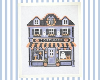 May include: A cross-stitch pattern on white fabric depicting a two-story building with the word COSTUMES printed on a sign. The design features purple, black, and orange colors, with ghost and pumpkin motifs, a striped awning, and windows.