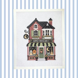 May include: A needlepoint canvas featuring a two-story building with the word APOTHECARY printed above the storefront. The design includes a black roof, red brick walls, green shutters, and windows displaying a pumpkin, moon, and broom.