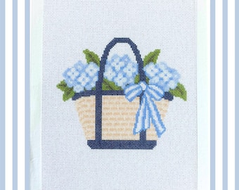 May include: A cross-stitch pattern on white fabric featuring a tan woven basket with a dark navy blue handle. The basket is filled with light blue hydrangea flowers and green leaves, accented by a light blue and white striped ribbon tied in a bow.