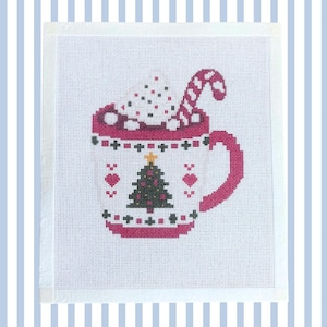 May include: A white cross-stitch fabric featuring a mug design in pink, dark green, and black. The mug contains a drink with whipped cream and a candy cane. A Christmas tree with a yellow star is centered on the mug, flanked by two pink hearts.