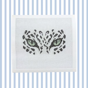 May include: A square needlepoint canvas featuring a cross-stitched design of a leopard's face. The pattern displays two piercing green eyes surrounded by black and grey spots on a white mesh background, creating a focused animal gaze.