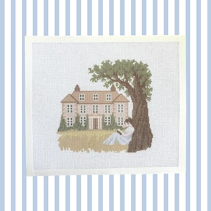 May include: A cross-stitch embroidery piece on white fabric featuring a large, multi-storey house with a tan facade and a brown roof. A large, dark brown tree stands to the right, with a person in a light blue outfit sitting underneath reading.