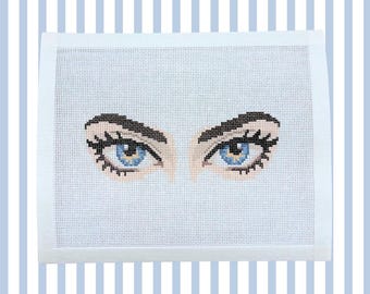 May include: A needlepoint canvas featuring a detailed cross-stitch design of two human eyes. The eyes have bright blue irises, dark brown eyebrows, and long black eyelashes, all rendered with precise stitching on a white grid fabric background.