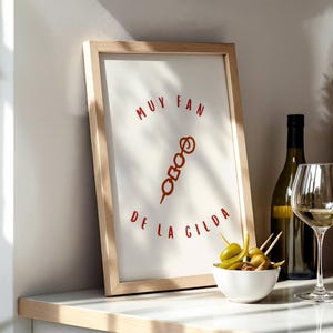 May include: A framed print with the words "MUY FAN DE LA GILDA" in red, featuring a skewer illustration. A bottle of wine, a wine glass, and a small bowl of olives with toothpicks are also visible.