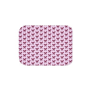 Puede incluir: Una alfombra rectangular con esquinas redondeadas, con un patrón repetitivo de lazos burdeos sobre un fondo rosa claro. Los lazos están espaciados uniformemente y detallados, creando un diseño visualmente atractivo. La alfombra probablemente está hecha de un material suave y duradero.