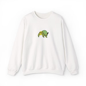 Puede incluir: Sudadera blanca de cuello redondo con un pequeño bordado de una tortuga de dibujos animados. La tortuga tiene una cara amarilla, ojos negros y un caparazón verde con detalles amarillos. La sudadera tiene mangas largas.